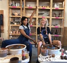 two women sitting on top of buckets in a pottery shop with one holding up her hand