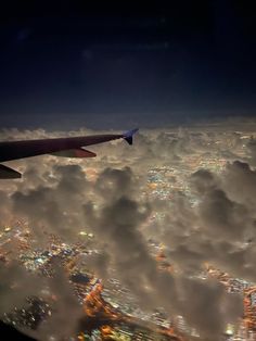 an airplane wing flying over the city lights in the night sky above some fluffy clouds