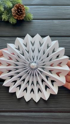 a hand holding a paper snowflake on top of a wooden table next to pine cones