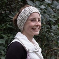 a smiling woman wearing a knitted headband in front of some trees and bushes