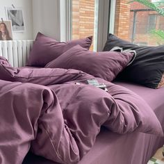 a bed covered in purple sheets and pillows next to a window with a brick wall