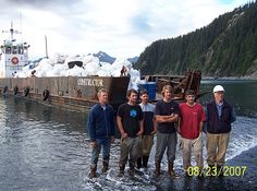 four men standing in the water next to a large boat that is being loaded with ice