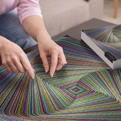 a woman is working on a colorful rug with her hands and two books in the background