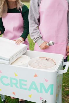 two women in aprons are making ice cream at an outdoor event, one is holding a box