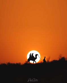 two people riding on the back of a horse in front of an orange sky at sunset