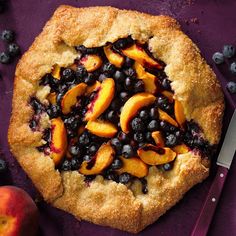 a pie with blueberries and peaches is on a purple surface next to a knife