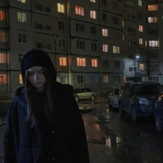 a woman standing in front of a building at night with cars parked on the street