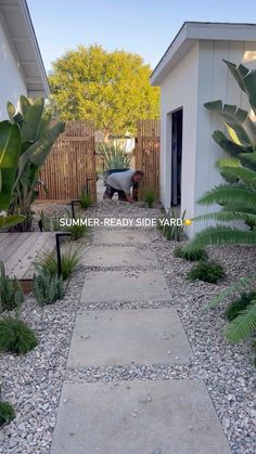 a walkway leading to a small house with plants in the foreground and a fenced yard behind it