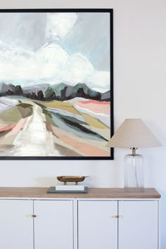 a painting hanging on the wall next to a white cabinet with a lamp in front of it