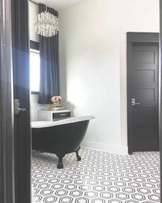 An elegant black and white modern minimalist bathroom showcases a large black clawfoot bathtub with modern black and white patterned tile bathroom flooring. White walls brighten the space and natural light from a window dressed with black curtains filters through the room. An intricate modern crystal chandelier is mounted over the tub and a small white cabinet rests in the corner...   Image: tx_desert_farmhouse