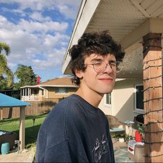 a young man wearing glasses standing in front of a house with a blue sky and clouds behind him