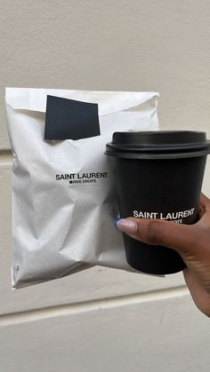 a hand holding a black coffee cup next to a white bag with saint laurent on it