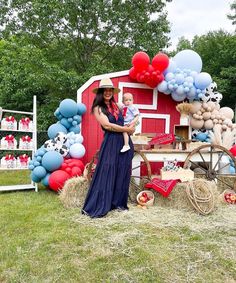 a woman holding a baby standing in front of a red barn with balloons and decorations