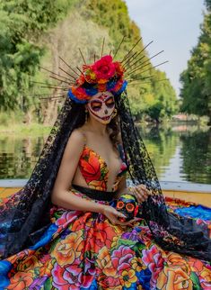 a woman in a colorful dress sitting on the edge of a body of water wearing a mask and veil