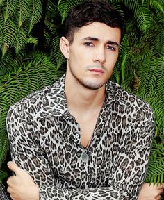 a man with his arms crossed standing in front of green plants and trees, wearing a leopard print shirt