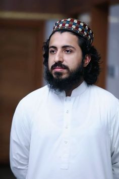 a man with a long beard wearing a white shirt and a headdress stands in front of a door