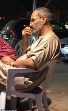 two men sitting at a table talking on their cell phones in front of parked cars