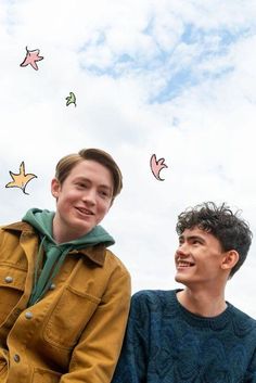 two young men sitting next to each other under a blue sky with kites flying above them