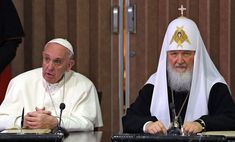 two men sitting at a table in front of microphones and one is wearing a priest's hat