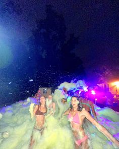 two women in bathing suits are playing in the water at night time with lights on behind them