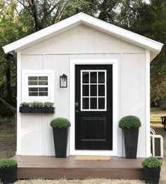 a small white shed with black doors and two planters on the front porch area