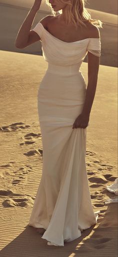 a woman standing in the sand wearing a white dress and holding her hair up to her face