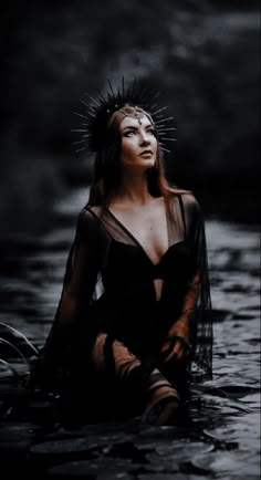 a woman sitting in the water wearing a sheer black dress and crown of spikes on her head