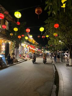 people are walking down the street at night with lanterns hanging above them and on motorcycles