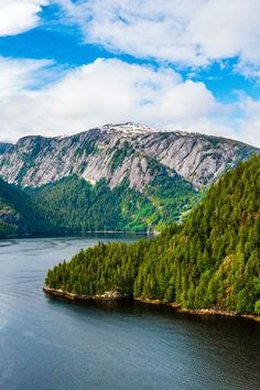 an island in the middle of a body of water surrounded by trees and mountains under a blue sky with white clouds