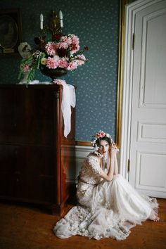 a woman in a wedding dress sitting on the floor next to a dresser with flowers