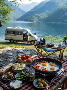 an outdoor table with food on it next to the water and a campervane