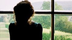 a woman standing in front of a window looking out at the grass and trees outside