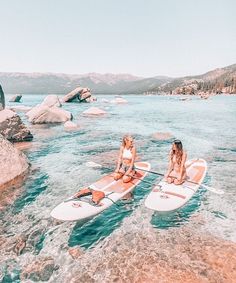 two women are sitting on surfboards in the water near rocks and boulders, while another woman is standing behind them