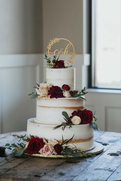 a three tiered wedding cake with red and white flowers on the top is sitting on a wooden table