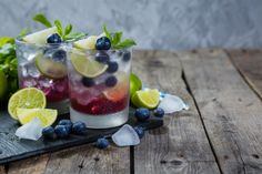 two glasses filled with blueberries, lemons and mint on top of a wooden table