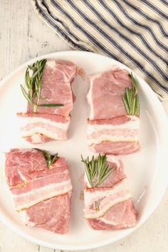 four pieces of raw meat on a white plate with rosemary sprigs and seasoning