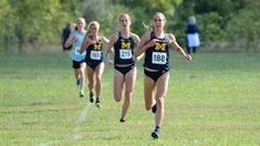 three girls running in a cross country race on the grass with trees in the background