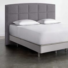 an upholstered bed with white pillows and gray headboard on concrete flooring