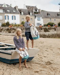 two young children standing on the beach next to a small boat with their name written on it