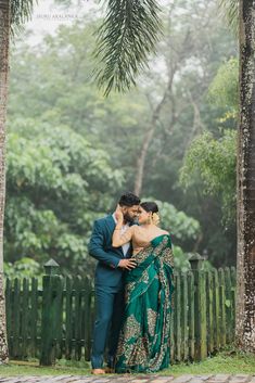 a man and woman standing next to each other in front of some trees with their arms around each other