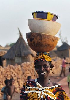 a woman with a large bowl on her head in front of some people and mud huts