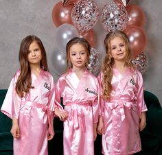 three girls in pink robes standing next to each other with balloons behind them and one girl wearing a name on her robe