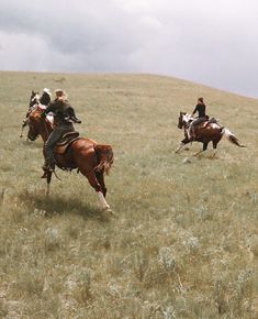 three people are riding horses in a grassy field on a cloudy day with dark clouds overhead