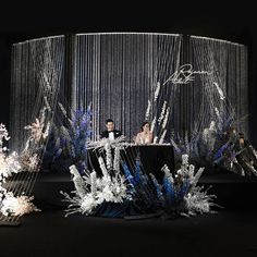 two people are sitting at a table surrounded by flowers and chains on the floor in front of a black backdrop