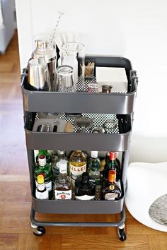 a bar cart filled with bottles and glasses on top of a hard wood floor next to a white wall