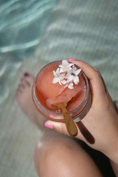 a woman holding a drink in her hand with flowers on the rim and water behind it