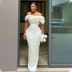 a woman in a white dress is standing by a door and holding a flower bouquet