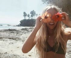 a woman standing on top of a sandy beach next to the ocean holding flowers in front of her eyes