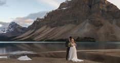 a bride and groom standing on the shore of a lake with mountains in the background