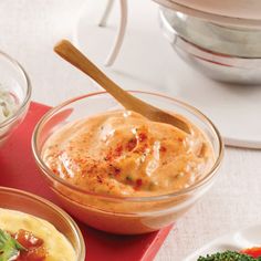 two bowls of dip and vegetables on a red place mat with a wooden spoon in the middle